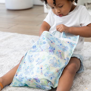 A girl sits on a gray rug, opening a bag from Bumkins The Chaos Coordinator Deluxe 5-Piece Set: Disney Aloha Stitch.