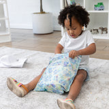 A young child with curly hair and a white outfit opens a colorful Bumkins Wet Bag: Aloha Stitch while sitting on the floor.