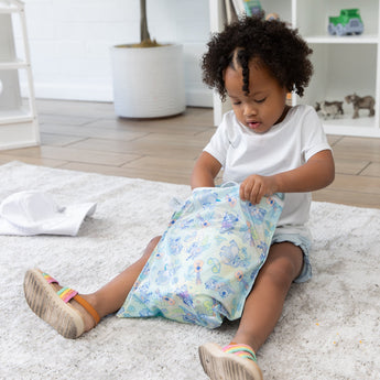 A young child with curly hair and a white outfit opens a colorful Bumkins Wet Bag: Aloha Stitch while sitting on the floor.