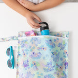 A child grabs a blue bottle from the Bumkins Wet Bag: Aloha Stitch on a white table with sunglasses nearby.