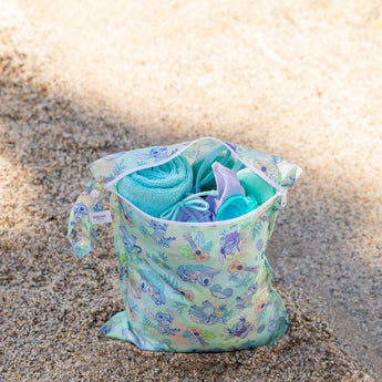 Bumkins Wet Bag: Aloha Stitch holds a towel and swimwear on a sandy beach shore.