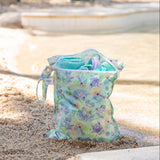 A bag from Bumkins Wet Bag + Wet/Dry Bag Set: Aloha Stitch sits on a sandy beach with a towel and swimwear inside.