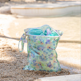 A bag from Bumkins Wet Bag + Wet/Dry Bag Set: Aloha Stitch sits on a sandy beach with a towel and swimwear inside.