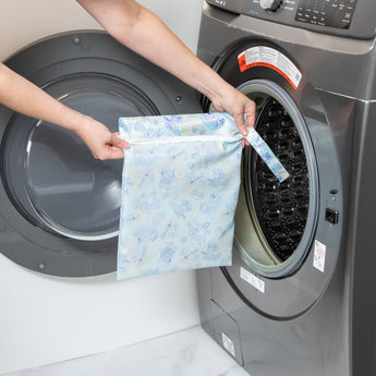 A person holds a Bumkins Wet Bag: Aloha Stitch in front of an open front-loading washing machine.
