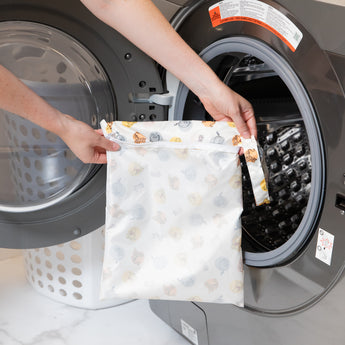 A person holds a bag from Bumkins The Chaos Coordinator Essential 4-Piece Set: Disney Winnie + Friends in front of an open washing machine.