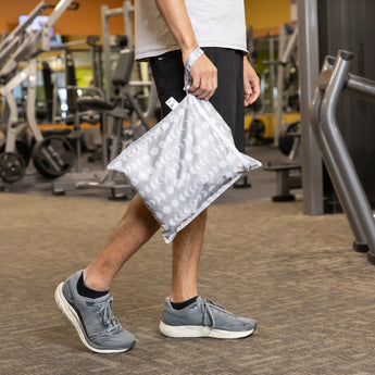 A gym-goer in athletic wear carries the Roamio by Bumkins Wet Bag 2 Pack: Desert Night past fitness equipment.