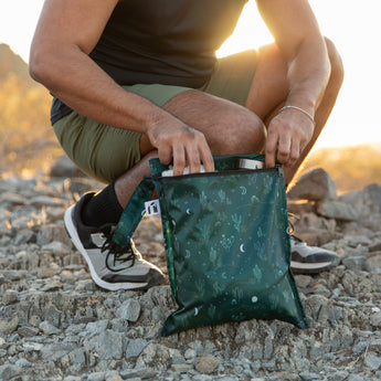 Kneeling outdoors, someone opens a Roamio by Bumkins Wet Bag 2 Pack: Desert Night, made from waterproof fabric.