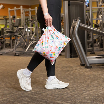 Person in gym clothes carries a Roamio by Bumkins Wet Bag 2 Pack: Disco Boots at the gym among exercise equipment.