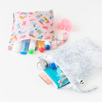 Roamio by Bumkins Wet Bag 2 Pack: Disco Boots lays open on a white background with toiletries, a book, glasses, & water bottle peeking out.