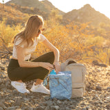 Woman kneels outdoors, packing a blue Wet Bag 2 Pack: Denim On Denim by Roamio into her backpack, dry hills behind.
