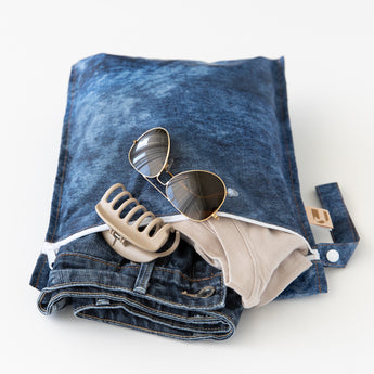 A Roamio by Bumkins Wet Bag 2 Pack: Denim On Denim rests open on a white background with clothes, sunglasses, & hair clip inside.