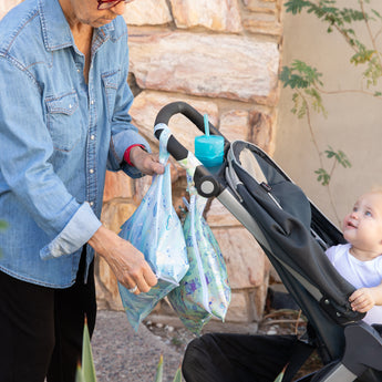An adult hangs Bumkins Wet Bag 2 Pack: Disney Stitch Star-ring Up Trouble on a stroller handle as a baby looks up.