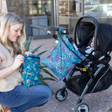 A woman kneels by stroller with a baby inside, zipping a Bumkins Wet Bag 2 Pack: Jungle + Animal Prints as another hangs from the handle.