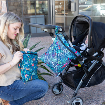 A woman kneels by stroller with a baby inside, zipping a Bumkins Wet Bag 2 Pack: Jungle + Animal Prints as another hangs from the handle.