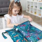 A young girl in a bow sits at a white table in front of a gray couch, reaching into Bumkins Wet Bag 2 Pack: Jungle + Animal Prints.