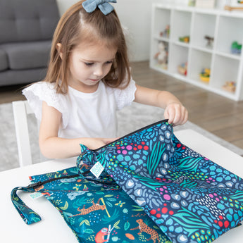 A young girl in a bow sits at a white table in front of a gray couch, reaching into Bumkins Wet Bag 2 Pack: Jungle + Animal Prints.
