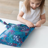 A child in a white shirt sits at a table, zipping a Bumkins Wet Bag 2 Pack: Jungle + Animal Prints.