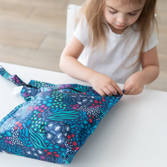 A child in a white shirt sits at a table, zipping a Bumkins Wet Bag 2 Pack: Jungle + Animal Prints.