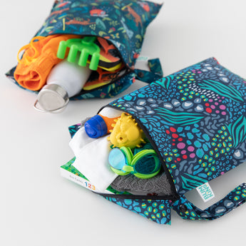 Bumkins Wet Bag 2 Pack: Jungle + Animal Prints, filled with beach essentials like goggles, a towel, and sunscreen, rests on a white background.