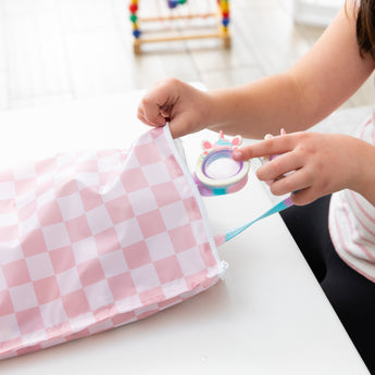 A child places goggles into a Bumkins Wet Bag Plus 2 Pack: Mauve + Sand Check on a white table.