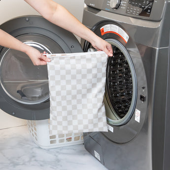 A person holds a Bumkins Wet Bag Plus 2 Pack: Mauve + Sand Check in front of an open front-loading washing machine.
