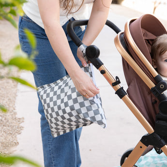 Outdoors, a woman unzips a Bumkins Wet Bag Plus 2 Pack: Mint + Charcoal Check attached to a stroller handle while a child sits inside.