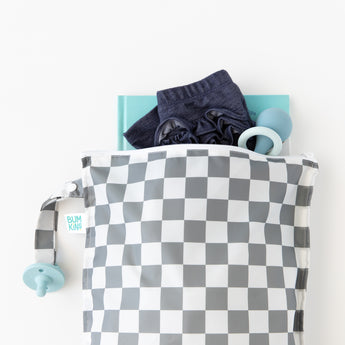 A Bumkins Wet Bag Plus 2 Pack: Mint + Charcoal Check holds baby items like a book, pants, teether, and pacifier on a white background.