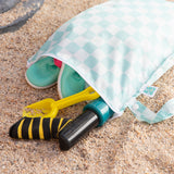 A Bumkins Wet Bag Plus 2 Pack: Mint + Charcoal Check lays open on sand with shoes and beach toys inside.