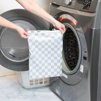 A person holds a Bumkins Wet Bag Plus 2 Pack: Mint + Charcoal Check in front of an open front-loading washing machine.