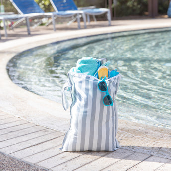 A Bumkins Wet Bag Plus 2 Pack: Green + Gray Stripe sits poolside holding sunglasses, sunscreen, a towel, and more.