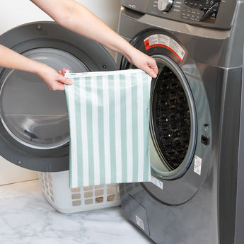 A person holds a Bumkins Wet Bag Plus 2 Pack: Green + Gray Stripe in front of an open front-loading washing machine.