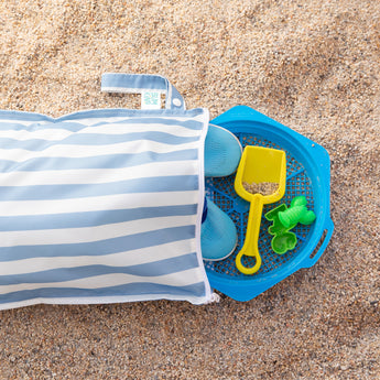 A Bumkins Wet Bag Plus 2 Pack: Blue + Yellow Stripe lays open on sand with shoes and beach toys spilling out.