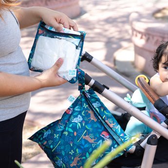 A person packs baby essentials in bags from Bumkins The Chaos Coordinator Essential 4-Piece Set: Jungle by a stroller with a baby outdoors.