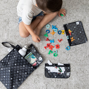 A boy plays with toys on gray carpet among bags from Bumkins The Chaos Coordinator Essential 4-Piece Set: Mickey Mouse Icon Black + White.