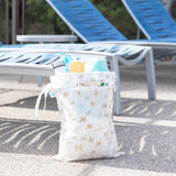 Bumkins Wet/Dry Bag: Palm Daze sits in front of empty lounge chairs, with a water bottle, towel, and swim gear inside.