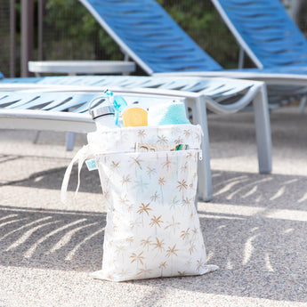 Bumkins Wet/Dry Bag: Palm Daze sits in front of empty lounge chairs, with a water bottle, towel, and swim gear inside.
