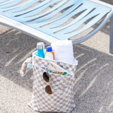 Bumkins Wet Bag + Wet/Dry Bag Set in Sand Check, packed with travel essentials, sits by a blue poolside lounge chair.