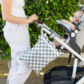 A woman pushes a stroller with a baby; a Bumkins Wet Bag + Wet/Dry Bag Set in Charcoal Check hangs from the handle.