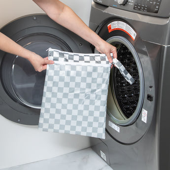 A person holds Bumkins Wet/Dry Bag: Charcoal Check in front of an open front-loading washing machine.
