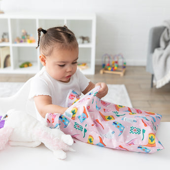 A girl reaches into a Bumkins Wet/Dry Bag: Boots at a white table next to a stuffed unicorn; toy filled shelves behind.