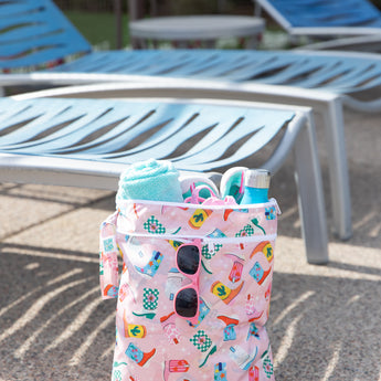 A Bumkins Wet Bag + Wet/Dry Bag Set: Boots holds sunglasses, a towel, swimwear, and a water bottle in front of empty blue lounge chairs.
