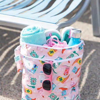 Bumkins Wet/Dry Bag: Boots holds a towel, water bottle, swimwear, and sunglasses in front of an empty lounge chair.