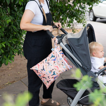 A person in black overalls pushes a stroller with a child, with a colorful Bumkins Wet/Dry Bag: Yoga Pals on the handle.