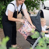 Someone zips a stylish Bumkins Wet/Dry Bag: Yoga Pals attached to a stroller with a child on a greenery-lined sidewalk.