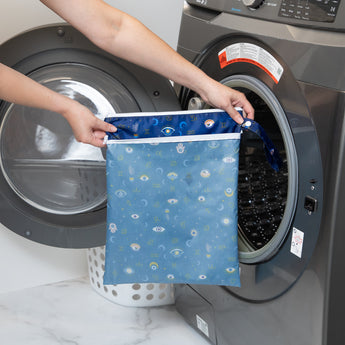 A person holds a bag from Bumkins Wet Bag + Wet/Dry Bag Set: Evil Eye in front of an open front-loading washing machine.