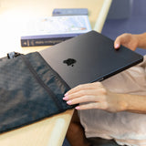 A person places a closed laptop into a Bumkins Wet/Dry Bag: Rich Black on a desk, next to a book.