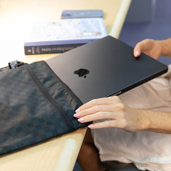 A person places a closed laptop into a Bumkins Wet/Dry Bag: Rich Black on a desk, next to a book.
