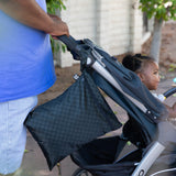A person in a blue shirt pushes a stroller with a Bumkins Wet/Dry Bag: Rich Black hanging from the handle, with trees in the background.