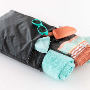 Bumkins Wet/Dry Bag: Rich Black holds a light blue towel, swim trunks, toy shovels, and teal sunglasses on a white background.