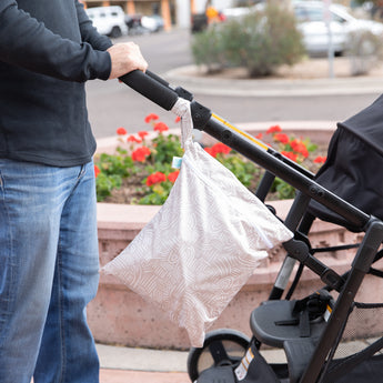 Someone pushes a stroller with Bumkins Wet/Dry Bag: Wander attached, surrounded by urban scenery and a flowerbed.
